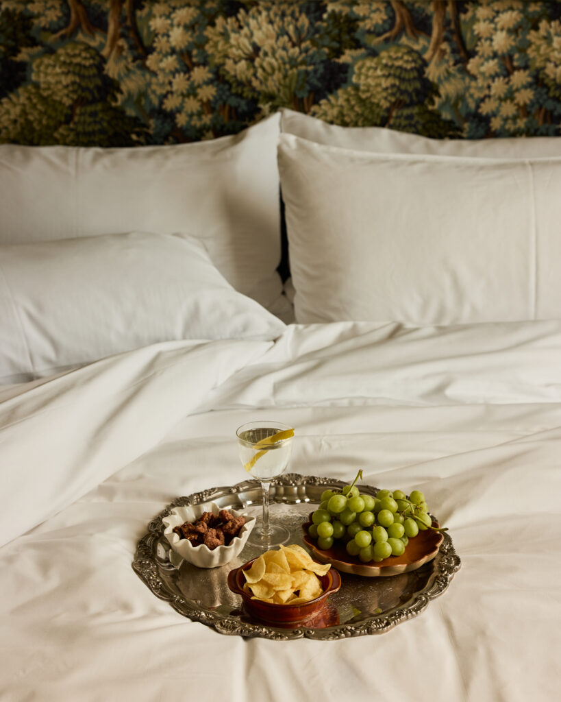 Room-service platter and a glass of wine on white bedding at The Nickel Hotel