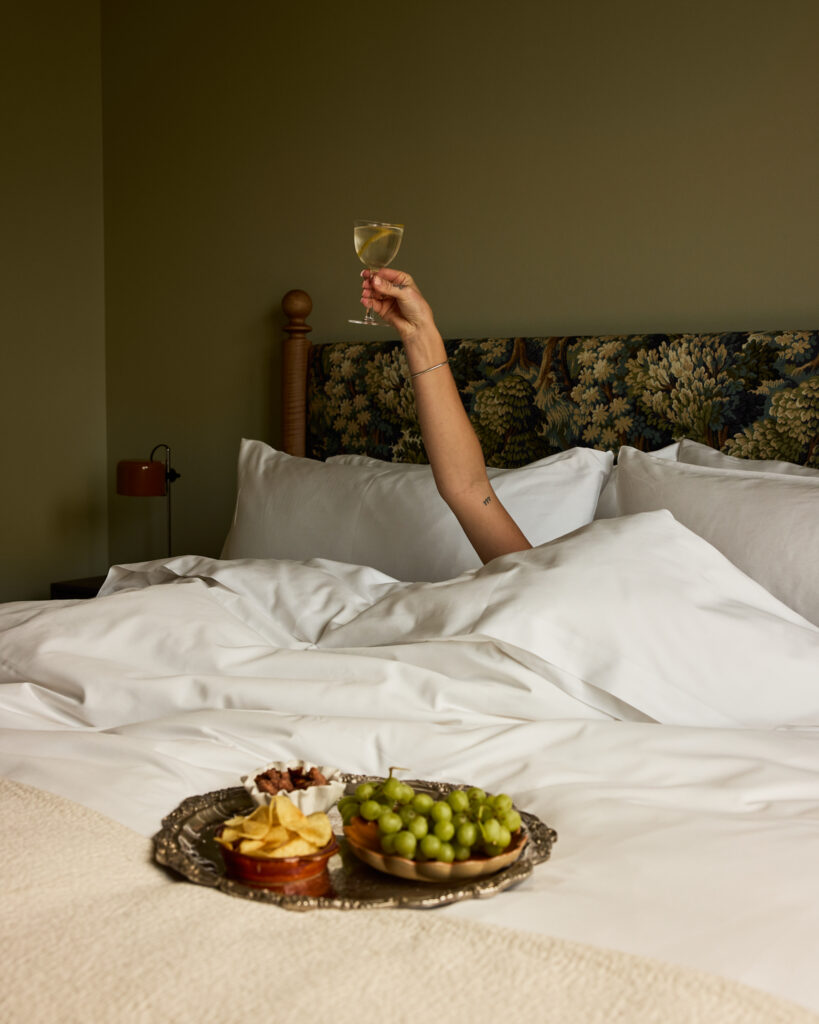 Guest raising a glass in bed beside a room-service platter at The Nickel Hotel